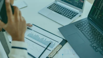 Na stole leżą dwa laptopy, na których widnieją wykresy. Mężczyzna siedzący przy tym stole rozmawia przez telefon zapoznając się z raportem