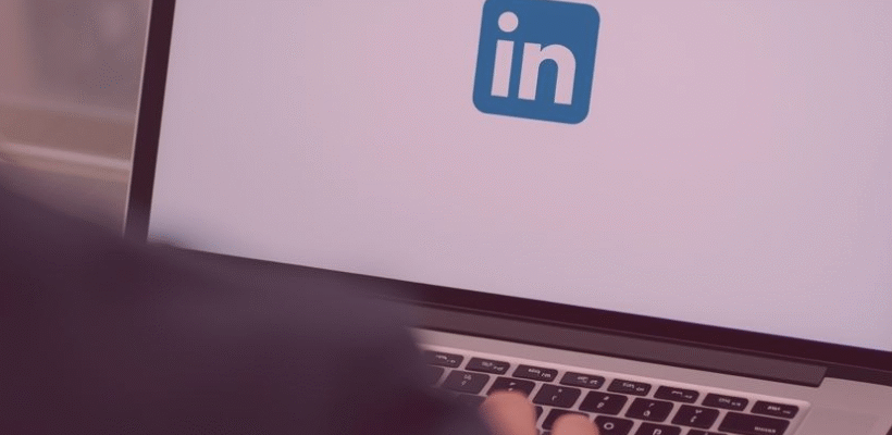 Na drewnianym stole stoi włączony laptop, na ekranie którego wyświetla się logo LinkedIn